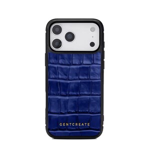 A crocodile-textured iPhone case. The case features a Glossy, reptile-inspired Glossy with raised scales that give it a luxurious, high-end look. The phone case has a black border, highlighting its sleek and modern design. The person's hand, with neatly manicured red nails, is seen against a backdrop of denim jeans, adding a stylish, fashionable touch to the image. The brand name "GENTCREATE" is subtly visible on the case.