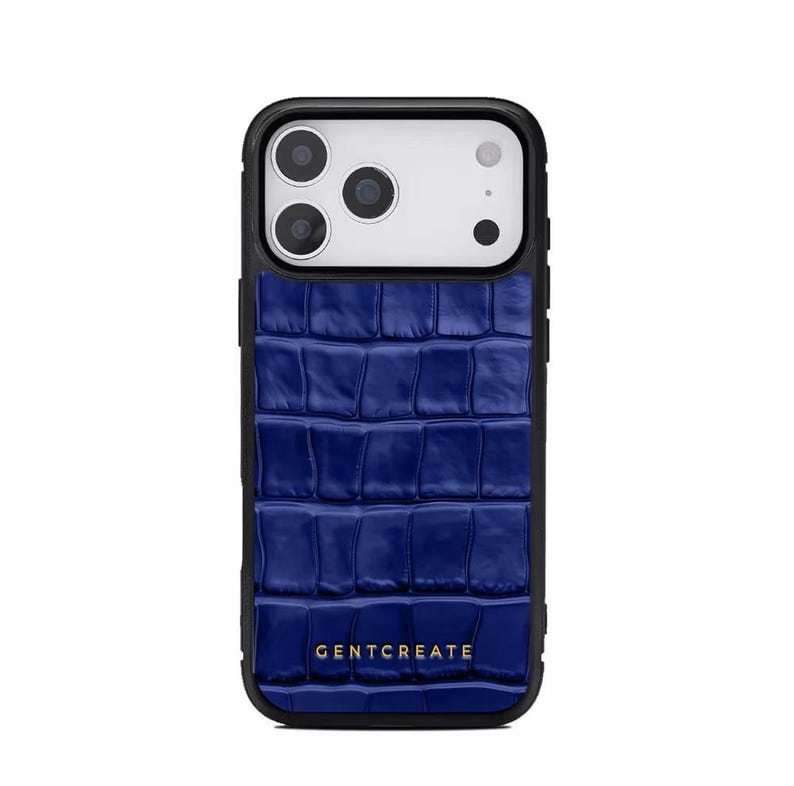 A crocodile-textured iPhone case. The case features a Glossy, reptile-inspired Glossy with raised scales that give it a luxurious, high-end look. The phone case has a black border, highlighting its sleek and modern design. The person's hand, with neatly manicured red nails, is seen against a backdrop of denim jeans, adding a stylish, fashionable touch to the image. The brand name "GENTCREATE" is subtly visible on the case.