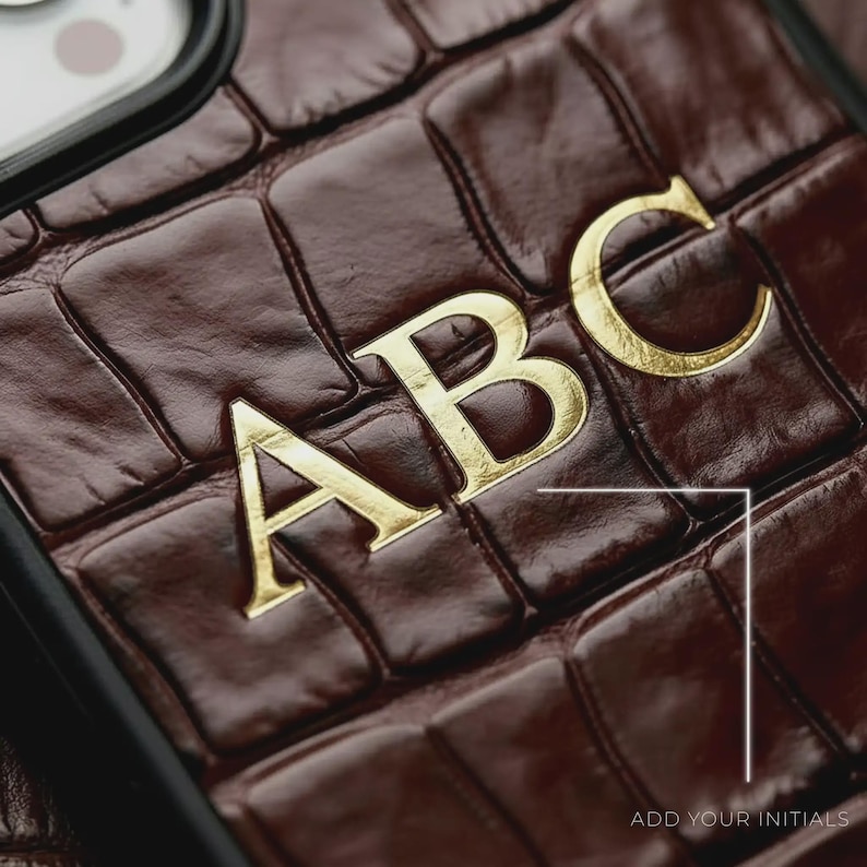 Puede incluir: Primer plano de una funda de tel&eacute;fono de cuero marr&oacute;n con un patr&oacute;n texturizado similar a la piel de cocodrilo. Las letras doradas "ABC" est&aacute;n adheridas a la funda. El texto "ADD YOUR INITIALS" es visible en la esquina inferior derecha.
