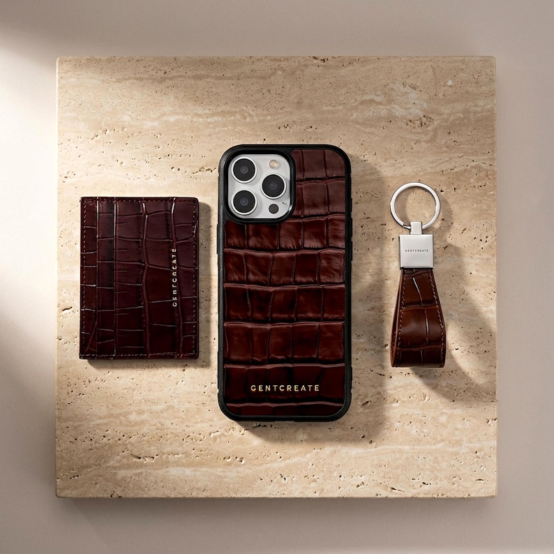 Puede incluir: Un conjunto de accesorios de cuero marr&oacute;n, que incluye una funda para tel&eacute;fono, un tarjetero y un llavero, dispuestos sobre una superficie texturizada. La funda del tel&eacute;fono y el tarjetero tienen una textura de piel de cocodrilo. El llavero tiene un anillo plateado.