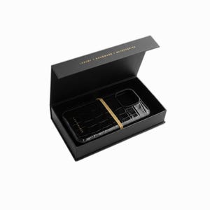 Puede incluir: Funda de tel&eacute;fono negra con textura de piel de cocodrilo, asegurada con una banda dorada, presentada en una caja negra. La tapa de la caja est&aacute; abierta, revelando la funda. El texto "LUXURY | HANDMADE | ACCESSORIES" est&aacute; impreso en la caja.