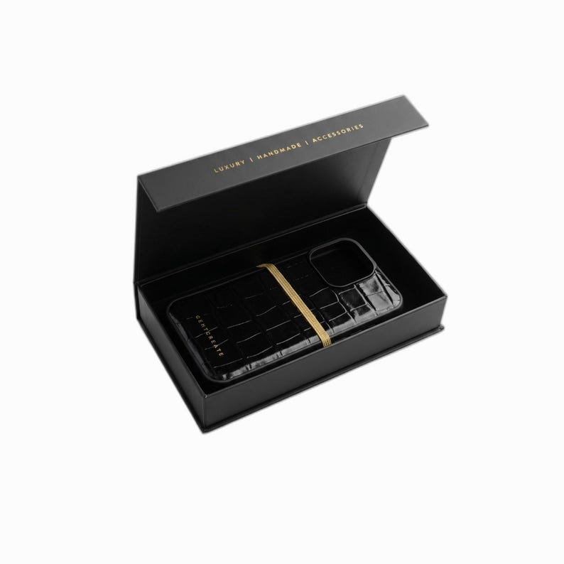 Puede incluir: Funda de tel&eacute;fono negra con textura de piel de cocodrilo, asegurada con una banda dorada, presentada en una caja negra. La tapa de la caja est&aacute; abierta, revelando la funda. El texto "LUXURY | HANDMADE | ACCESSORIES" est&aacute; impreso en la caja.