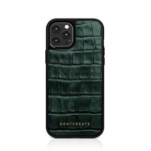 A crocodile-textured iPhone case. The case features a Glossy, reptile-inspired Glossy with raised scales that give it a luxurious, high-end look. The phone case has a black border, highlighting its sleek and modern design. The person's hand, with neatly manicured red nails, is seen against a backdrop of denim jeans, adding a stylish, fashionable touch to the image. The brand name "GENTCREATE" is subtly visible on the case.