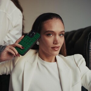 Puede incluir: Se muestra una funda de tel&eacute;fono verde con textura de piel de cocodrilo. La funda tiene un borde negro y cubre la c&aacute;mara del tel&eacute;fono. La persona de la imagen lleva una blazer blanca y una camisa blanca.