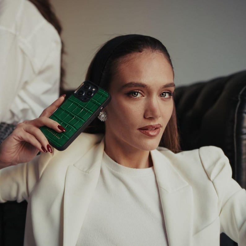 Puede incluir: Se muestra una funda de tel&eacute;fono verde con textura de piel de cocodrilo. La funda tiene un borde negro y cubre la c&aacute;mara del tel&eacute;fono. La persona de la imagen lleva una blazer blanca y una camisa blanca.
