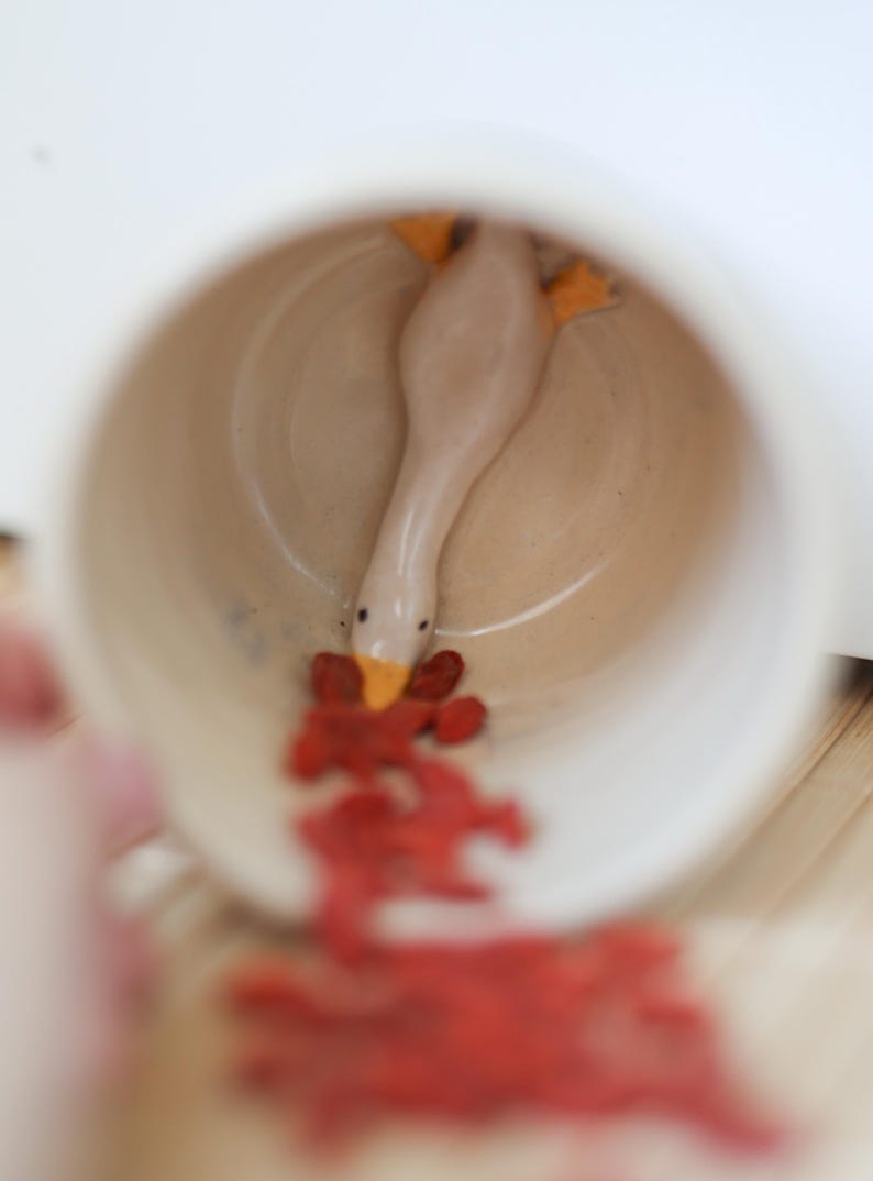 Puede incluir: Una taza de cer&aacute;mica blanca con una peque&ntilde;a figurita de ganso de cer&aacute;mica en su interior. El ganso est&aacute; sacando la cabeza de la taza y tiene el pico abierto. Hay bayas de goji rojas esparcidas alrededor de la taza.