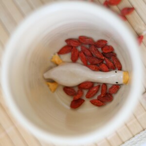 Puede incluir: Una figurita de ganso de cer&aacute;mica blanca con pico y patas amarillas se encuentra dentro de una taza de cer&aacute;mica blanca. La taza est&aacute; llena de bayas de goji rojas.