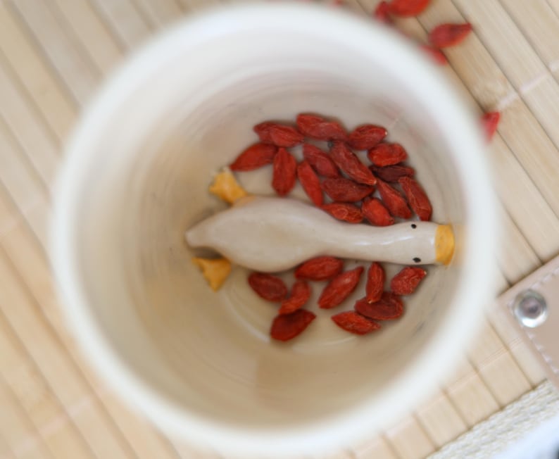 Puede incluir: Una figurita de ganso de cer&aacute;mica blanca con pico y patas amarillas se encuentra dentro de una taza de cer&aacute;mica blanca. La taza est&aacute; llena de bayas de goji rojas.