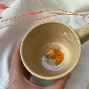 Peut inclure: Une tasse en céramique beige avec un glaçage moucheté et une anse brune. À l'intérieur de la tasse se trouve une petite figurine de renard en céramique peinte en orange et jaune.
