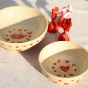 May include: Two white ceramic bowls with red heart and love designs. The bowls are on a white linen tablecloth.