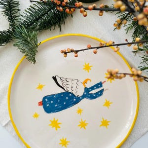 Op de afbeelding: Witte keramische plaat met gele rand. De plaat heeft een handgeschilderde illustratie van een vliegende engel in een blauwe jurk met witte vleugels en gele sterren. Perfect om eten op te serveren of als decoratief stuk.