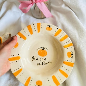 May include: A white ceramic plate with orange pumpkin and stripe designs. The plate has the words "Happy autumn" written in black script. The plate is decorated with orange pumpkins and stripes.