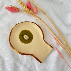 May include: A handmade ceramic dish with a kiwi slice center. The dish is off-white with a brown rim and a unique shape. Dried wheat stalks and pink flowers are arranged nearby, creating a natural, decorative display.