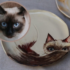 May include: A white ceramic plate featuring a hand-painted design of a Siamese cat. The cat is depicted in brown, black, and white, with blue eyes. This plate is a unique and decorative item for any cat enthusiast.