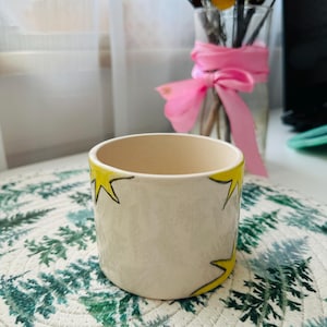 Op de afbeelding: Een kleine, crèmekleurige keramische pot met een gele sterrenprint. De pot heeft een cilindrische vorm en een gladde, ongeglazuurde buitenkant. De sterren zijn zwart omlijnd. De pot staat op een groen-witte placemat.
