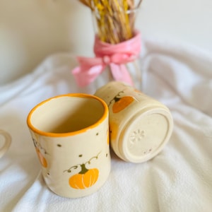 Op de afbeelding: Twee keramische mokken met een crèmekleurige basis en oranje randen. Elke mok heeft een handgeschilderd oranje pompoenontwerp met groene ranken en kleine zwarte stippen. De mokken staan op een wit oppervlak, wat wijst op een herfstthema.