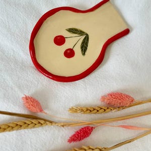 Könnte beinhalten: Ein Keramik-Löffelablage mit cremefarbener Oberfläche und rotem Rand. Die Löffelablage zeigt ein Kirschdesign mit zwei roten Kirschen und grünen Blättern. Getrocknete Blumen und Weizenähren befinden sich im Hintergrund.