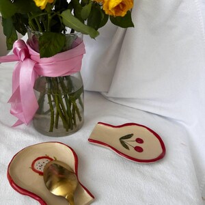 Könnte beinhalten: Eine klare Glasvase mit gelben Rosen und einem rosa Band. Zwei Keramik-Löffelablagen mit rotem Rand und Fruchtdesign auf weißer Oberfläche. Eine Löffelablage mit goldenem Löffel.