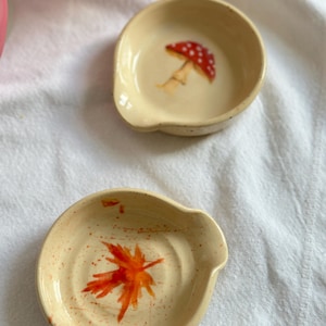 Può includere: Due piccoli poggiacucchiai rotondi in ceramica. Uno presenta un fungo rosso a pois bianchi, l'altro una foglia d'acero arancione stilizzata. Entrambi sono beige con macchie arancioni e un piccolo beccuccio.