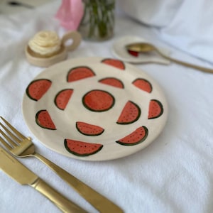 Può includere: Un piatto in ceramica bianca decorato con fette di anguria rosse e un centro rosso. Una forchetta e un coltello dorati sono a sinistra. Un piattino con un cucchiaio dorato e un piattino con burro sono sullo sfondo.