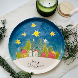 Op de afbeelding: Een handbeschilderde keramische plaat met een winterdorpsscène. De plaat heeft een blauwe lucht met gele sterren, een witte met sneeuw bedekte basis en kleurrijke huizen en bomen. De woorden "Merry Christmass" staan op de plaat.