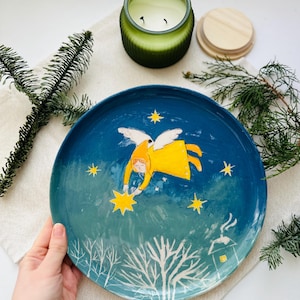 Op de afbeelding: Een handbeschilderde keramische plaat met een hemels thema. De plaat heeft een blauwe en blauwgroene achtergrond met een geel geklede engel die naar een ster reikt. Witte bomen en een klein huis staan ​​onderaan. Een groene kaars en dennentakken staan ​​op de achtergrond.