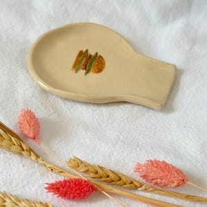 Puede incluir: Un reposacucharas de cerámica beige con un diseño de sándwich pintado. El reposacucharas tiene forma ovalada con un asa. Espigas de trigo secas y flores rosas están dispuestas alrededor del reposacucharas sobre una superficie blanca.