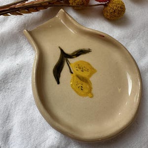 Könnte beinhalten: Ein Keramik-Löffelablage in Tropfenform mit handbemaltem Design aus zwei gelben Zitronen an einem Zweig mit grünen Blättern.