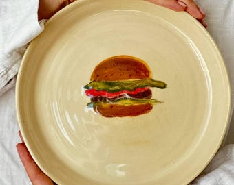 May include: A round, cream-colored ceramic plate featuring a hand-painted burger design. The burger has a brown bun, red tomato, green lettuce, and a yellow patty. The plate is held by a person wearing a white shirt with red heart accents.