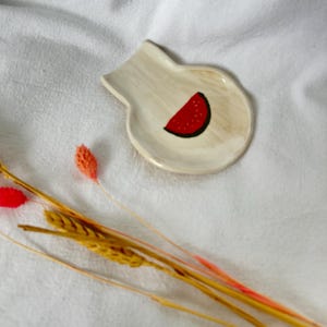 Puede incluir: Un pequeño plato de cerámica blanquecina con un diseño de rodaja de sandía. La sandía es roja con semillas negras y un contorno negro. El plato tiene una forma única y está colocado sobre una superficie blanca texturizada. Flores secas en primer plano.