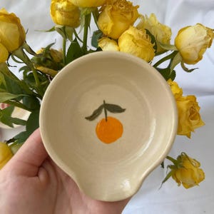 Pode incluir: Um pequeno descanso de colher de cerâmica esbranquiçada com um desenho pintado à mão de uma laranja e uma folha verde. O descanso de colher é redondo com uma pega curva. Rosas amarelas no fundo.