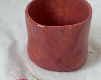 May include: A handmade ceramic cup with a textured, pink glaze. The cup has a rounded shape and a wide opening.