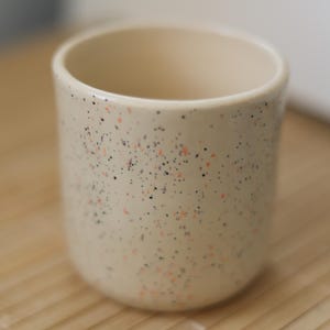 Puede incluir: Una taza de cer&aacute;mica beige moteada con motas naranja y negras. La taza est&aacute; sobre una estera de bamb&uacute; tejida.