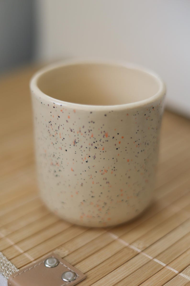 Puede incluir: Una taza de cer&aacute;mica beige moteada con motas naranja y negras. La taza est&aacute; sobre una estera de bamb&uacute; tejida.
