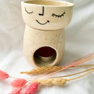 Op de afbeelding: Een crèmekleurige keramische etherische olie diffuser met een lachend gezichtsontwerp. De diffuser heeft een ronde bovenkant en een basis met een cirkelvormige opening voor een waxinelichtje. Roze en beige gedroogde bloemen staan op de voorgrond.