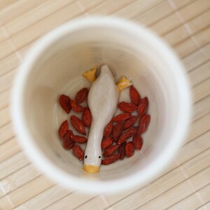 Puede incluir: Una taza de cer&aacute;mica blanca con una peque&ntilde;a figura de ganso de cer&aacute;mica en su interior. El ganso est&aacute; acostado de espaldas con las patas en el aire. La taza est&aacute; llena de bayas de goji rojas.