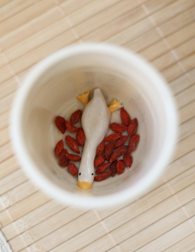 Puede incluir: Una taza de cer&aacute;mica blanca con una peque&ntilde;a figura de ganso de cer&aacute;mica en su interior. El ganso est&aacute; acostado de espaldas con las patas en el aire. La taza est&aacute; llena de bayas de goji rojas.