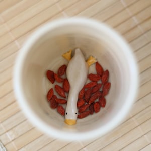 May include: A white ceramic cup with a small ceramic duck figurine inside. The duck is facing down with its head and beak visible. The cup is filled with red goji berries.