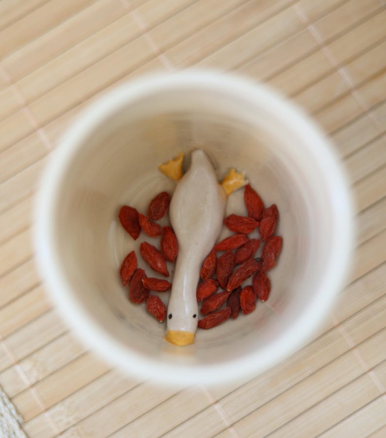 Puede incluir: Una taza de cer&aacute;mica blanca con una peque&ntilde;a figura de pato de cer&aacute;mica en su interior. El pato est&aacute; boca abajo con la cabeza y el pico visibles. La taza est&aacute; llena de bayas de goji rojas.