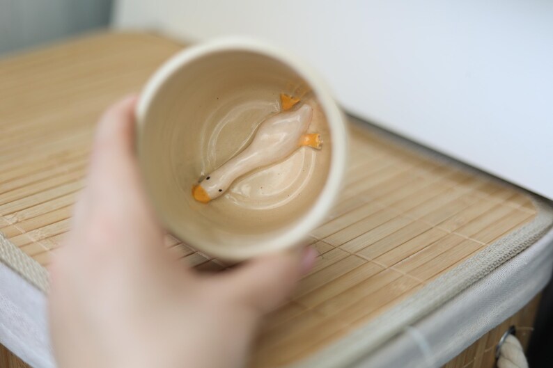 Puede incluir: Una taza de cer&aacute;mica blanca con una peque&ntilde;a figura de pato blanco en su interior. El pato tiene patas y un pico de color naranja.