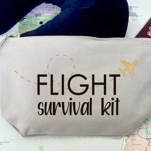 May include: A light gray zippered pouch with the text "FLIGHT survival kit" in black lettering. A gold airplane icon is on the right side of the pouch. The pouch is on a map with a passport.