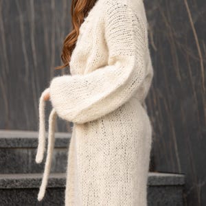 May include: A long-sleeved, off-white cardigan made of a chunky, loose-knit yarn. The garment features a relaxed fit with dropped shoulders, voluminous sleeves, and a matching tie belt that hangs down from the waist area, creating a soft texture.