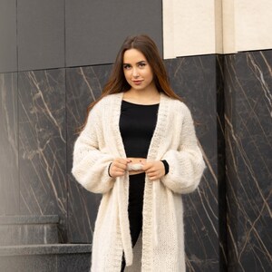 May include: A long, cream-colored, open-front knit cardigan featuring a chunky, fuzzy texture, long sleeves with ribbed cuffs, and a matching tie belt. The garment is worn over a black crop top and black trousers, creating a layered look.