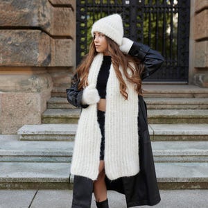 Puede incluir: Un gorro de punto blanco esponjoso, una bufanda larga de punto blanco esponjoso a juego y manoplas de punto blanco esponjoso. El conjunto se lleva sobre un top corto negro y un abrigo largo de cuero negro. Los accesorios tienen una textura suave.
