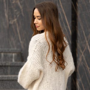 May include: An off-white, chunky knit sweater featuring a loose, open-weave texture with long, voluminous sleeves. The garment has a relaxed fit and a soft, fuzzy appearance, providing a cozy look while maintaining a simple, clean design.