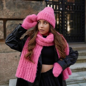 Puede incluir: Un gorro de punto rosa brillante, una bufanda larga de canal&eacute; a juego y un par de manoplas de punto a juego. El conjunto presenta una construcci&oacute;n de hilo difuso y texturizado en un tono rosa vibrante, ofreciendo un aspecto coherente.