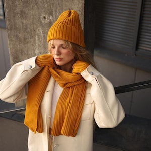 Könnte beinhalten: Eine Frau trägt einen weißen Mantel mit einer senfgelben Strickmütze, einem Schal und Handschuhen.