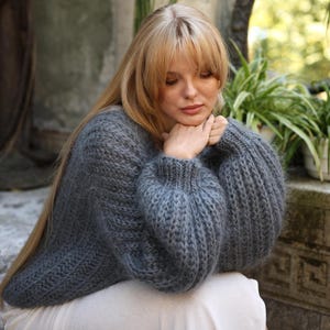Peut inclure: Un pull en tricot épais de couleur gris anthracite foncé présentant une composition en fil texturé et duveteux, une coupe décontractée et des manches longues volumineuses avec des poignets côtelés. Le vêtement a un col rond.