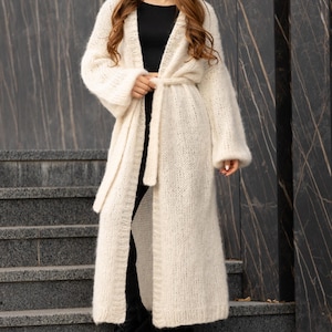 May include: A long, cream-colored, open-front knit cardigan featuring a fuzzy texture, long sleeves, and a matching tie belt at the waist. The garment is worn over a simple black top and black pants, extending down to the lower calf area.