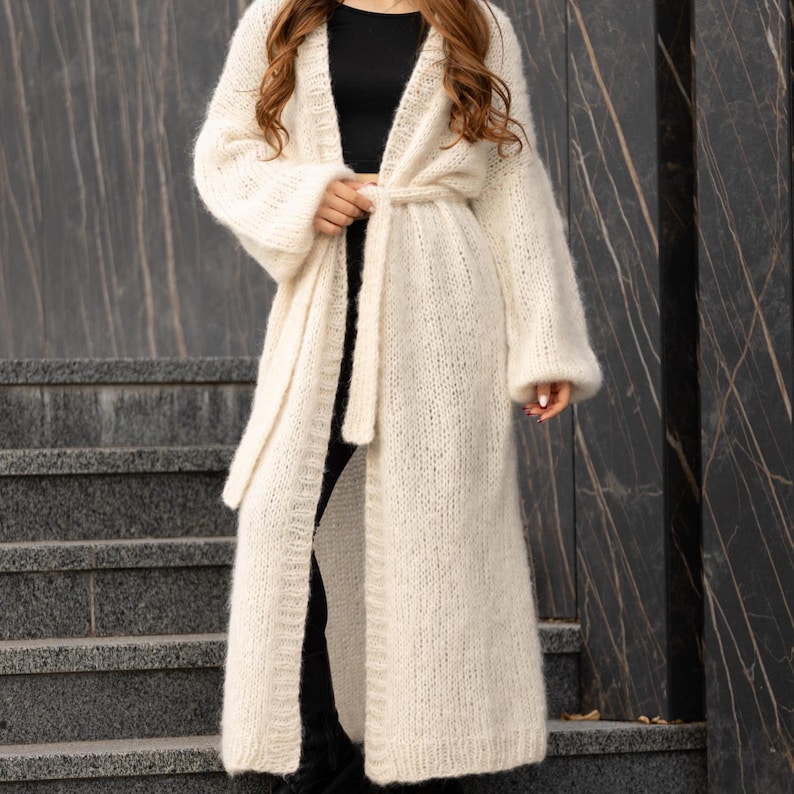 May include: A long, cream-colored, open-front knit cardigan featuring a fuzzy texture, long sleeves, and a matching tie belt at the waist. The garment is worn over a simple black top and black pants, extending down to the lower calf area.